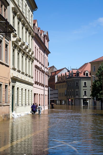 A scene of urban flooding in a residential area with several multi-story buildings, some with decorative window trims, adjacent to a street submerged in water. The water covers the pavement and extend