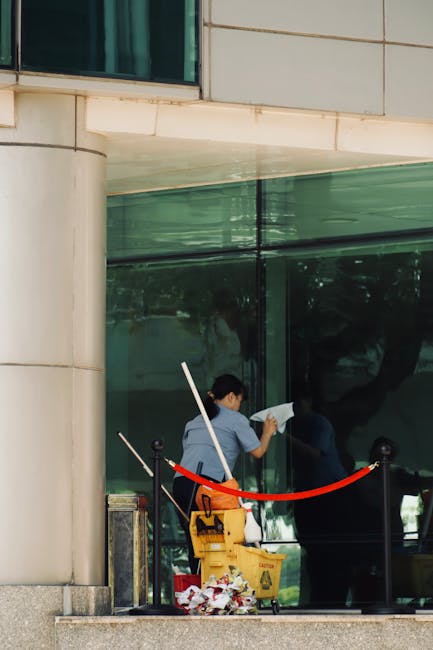 A person wearing a beige jumpsuit is performing surface cleaning with a vacuum cleaner on a dark carpeted floor in a commercial space. The room features a black wall with a white panel above, and a sm