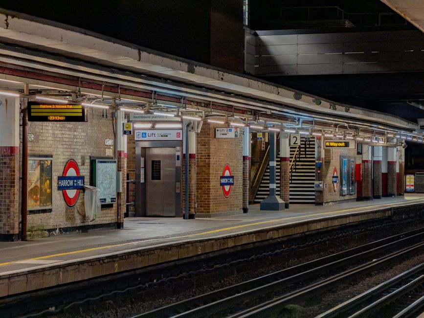A view of Wealdstone Station platform at night, featuring brick walls with signage indicating Harrow and Wealdstone station and directions for lifts and exit. The platform surface is clean, with a yel